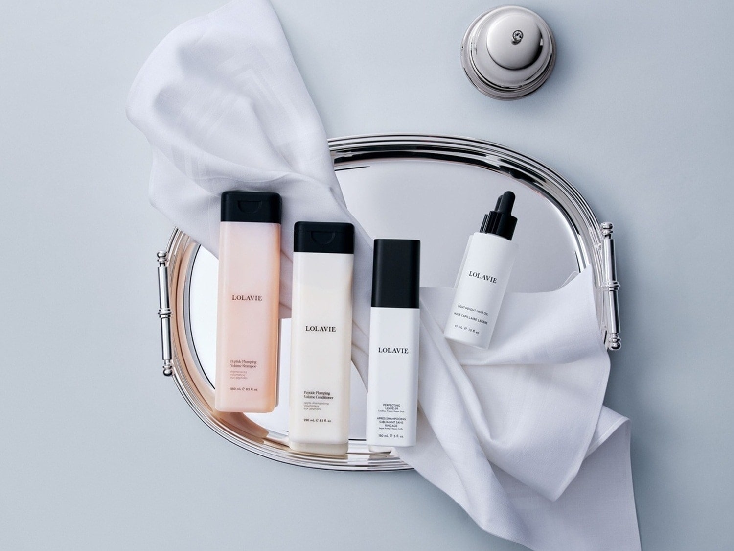 Descubre Lolavie: champú voluminizador en envase rosa, acondicionador en blanco, aceite capilar ligero y leave-in perfectos sobre una bandeja plateada con una toalla blanca.