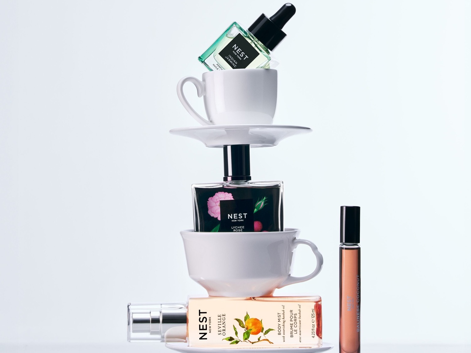 En la imagen se aprecia una torre de tazas de té blancas que sostienen varios productos de belleza de NEST, incluyendo aceites perfumados, brumas corporales y perfumes.