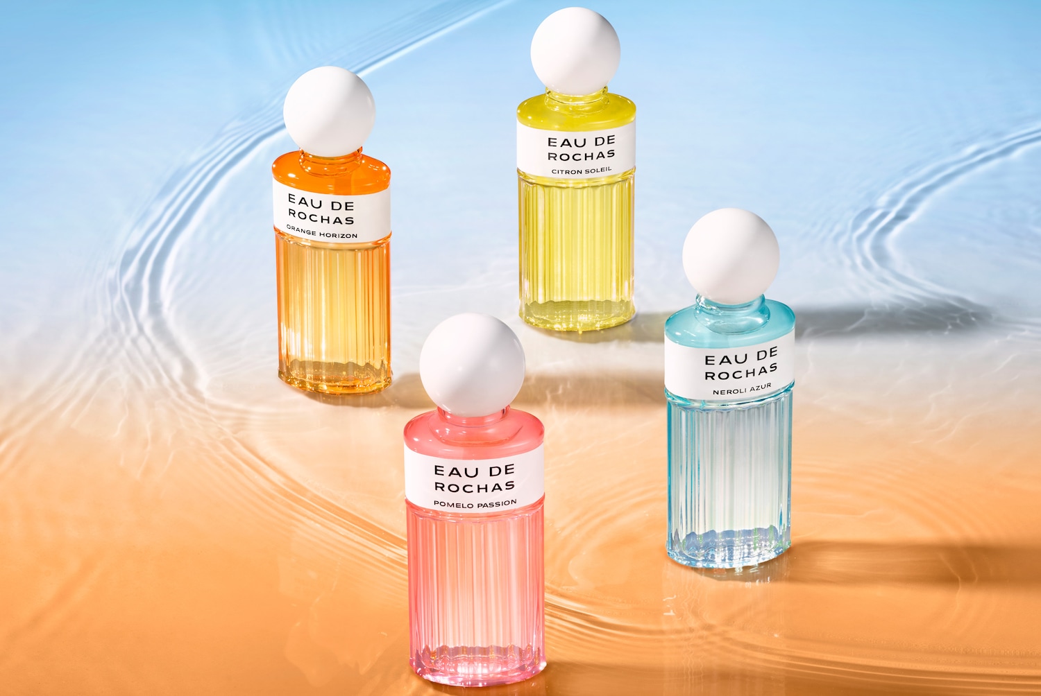 Quatre bouteilles de parfum Eau de Rochas aux couleurs vives : orange horizon, pomelo passion, citron soleil et néroli azur, chacune avec un bouchon blanc arrondi.