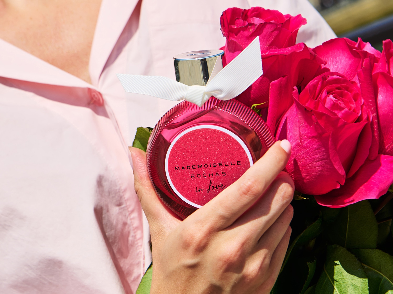 Une personne tenant un parfum Mademoiselle Rochas In Love rose vif, orné d'un ruban blanc, entouré de roses rouges vibrantes.