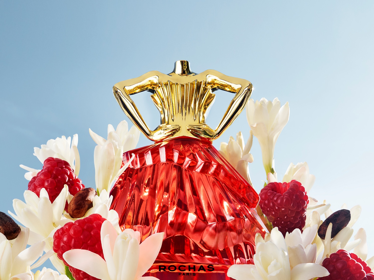 Découvrez Rochas Paris, un parfum dans un flacon rouge en forme de robe, orné d'un bouchon doré, entouré de fleurs blanches, de framboises et de fèves de cacao.