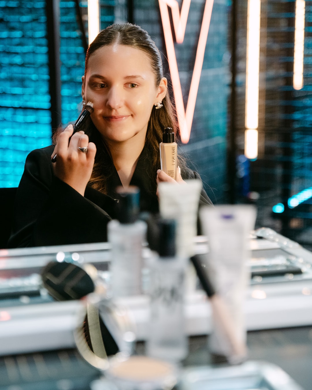 Een jonge vrouw brengt foundation aan met een kwast, terwijl ze een fles Lancôme foundation vasthoudt, omringd door andere make-upproducten.
