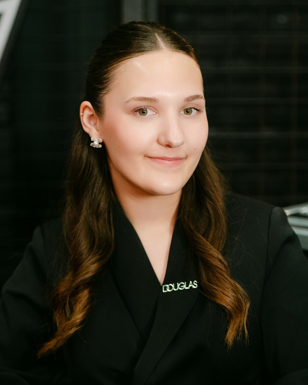 Portret van een jonge vrouw met bruin haar, gekleed in een zwarte blazer met een Douglas-badge, lachend naar de camera.