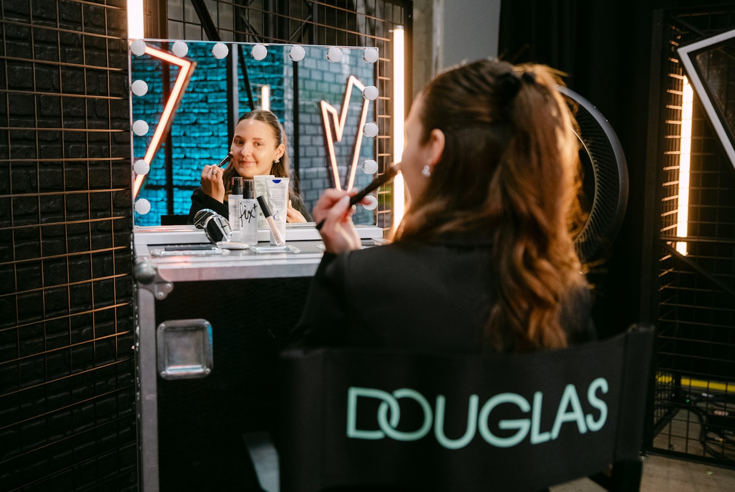 Een vrouw zit voor een spiegel met lampjes en brengt make-up aan met een kwast, terwijl ze op een regisseursstoel zit met het Douglas-logo erop.