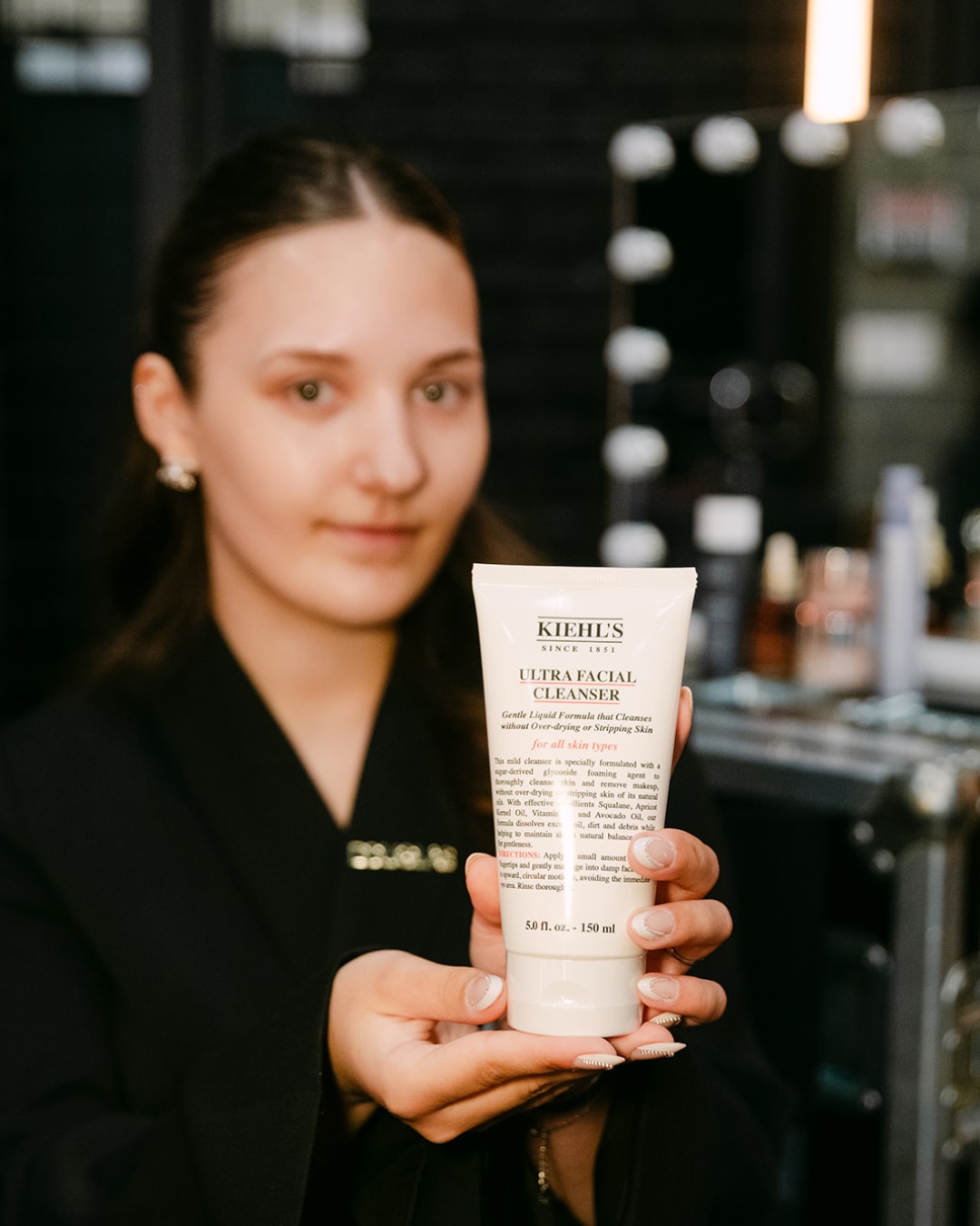 Een vrouw toont Kiehl's Ultra Facial Cleanser in een witte tube, die een zachte formule belooft voor alle huidtypes, reinigt zonder uit te drogen.