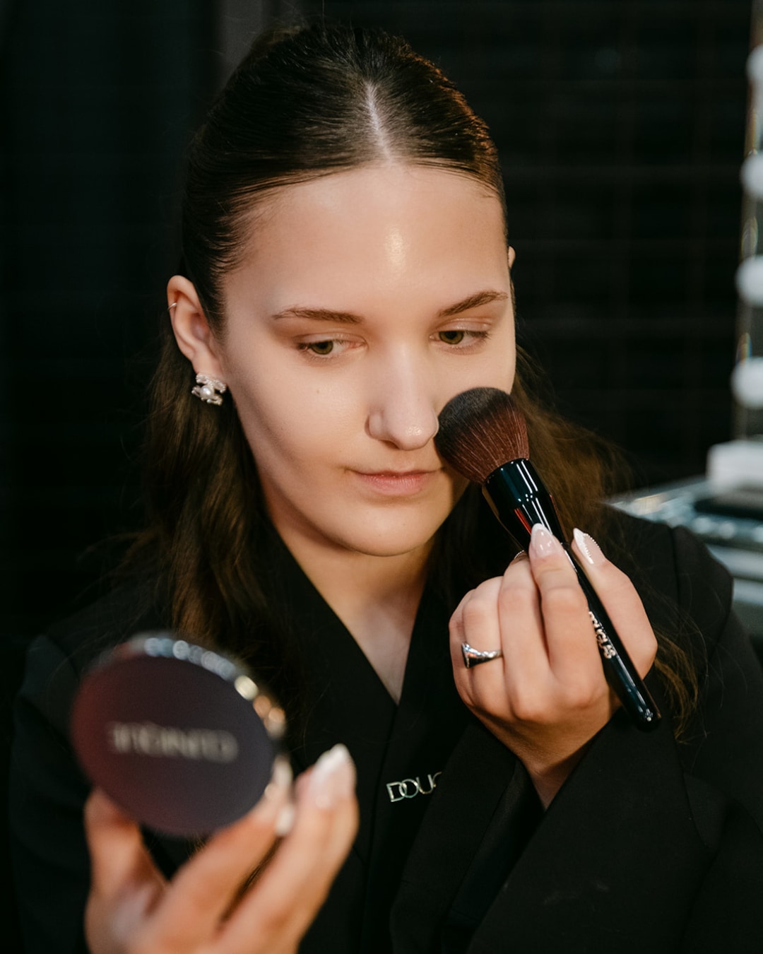 Een jonge vrouw brengt make-up aan met een grote, zachte borstel, terwijl ze in een compacte spiegel kijkt, gekleed in een zwarte kimono-stijl jas.
