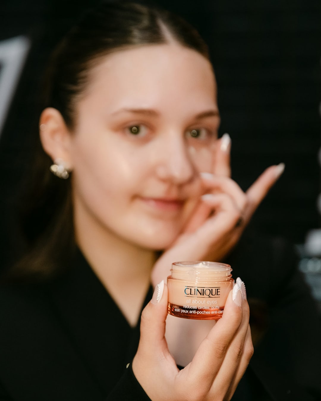 Een vrouw houdt een potje Clinique All About Eyes oogcrème vast, die kringen en wallen vermindert, terwijl ze de crème aanbrengt.
