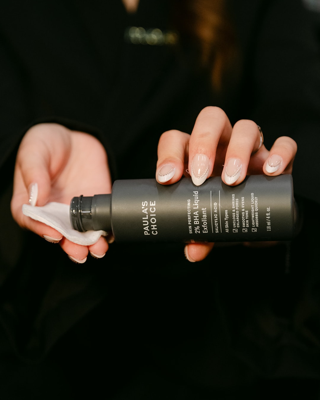 Een grijze fles Paula's Choice 2% BHA Liquid Exfoliant wordt vastgehouden, terwijl een vrouw het product op een wattenschijfje aanbrengt, ideaal voor het ontstoppen van poriën en het egaliseren van de huid.