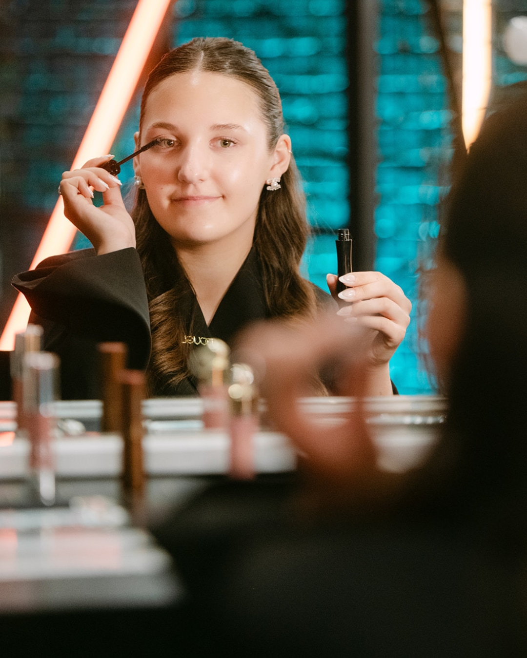 Een jonge vrouw brengt zwarte mascara aan op haar wimpers, kijkend in een spiegel met make-upproducten op de voorgrond.