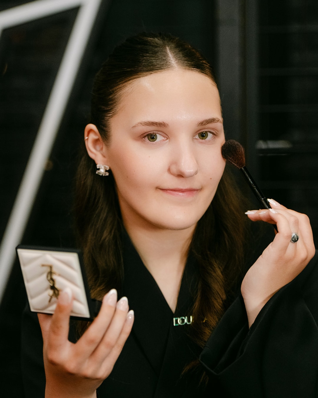 Een jonge vrouw brengt make-up aan met een kwast en een YSL-compactpoederdoos in haar hand.