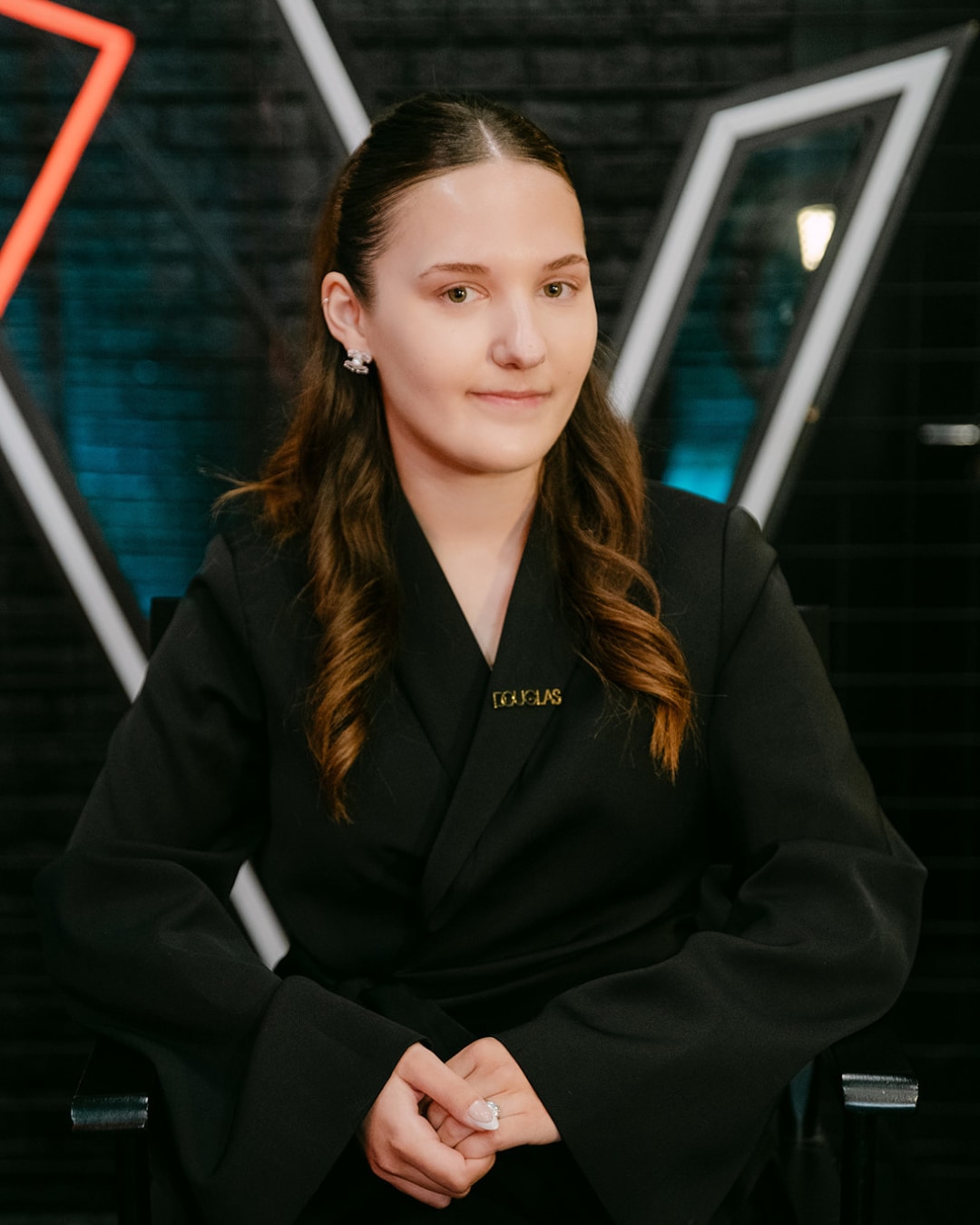 Een jonge vrouw met bruin haar en een zwarte blazer met een Douglas-speldje, zit voor een achtergrond met geometrische vormen.