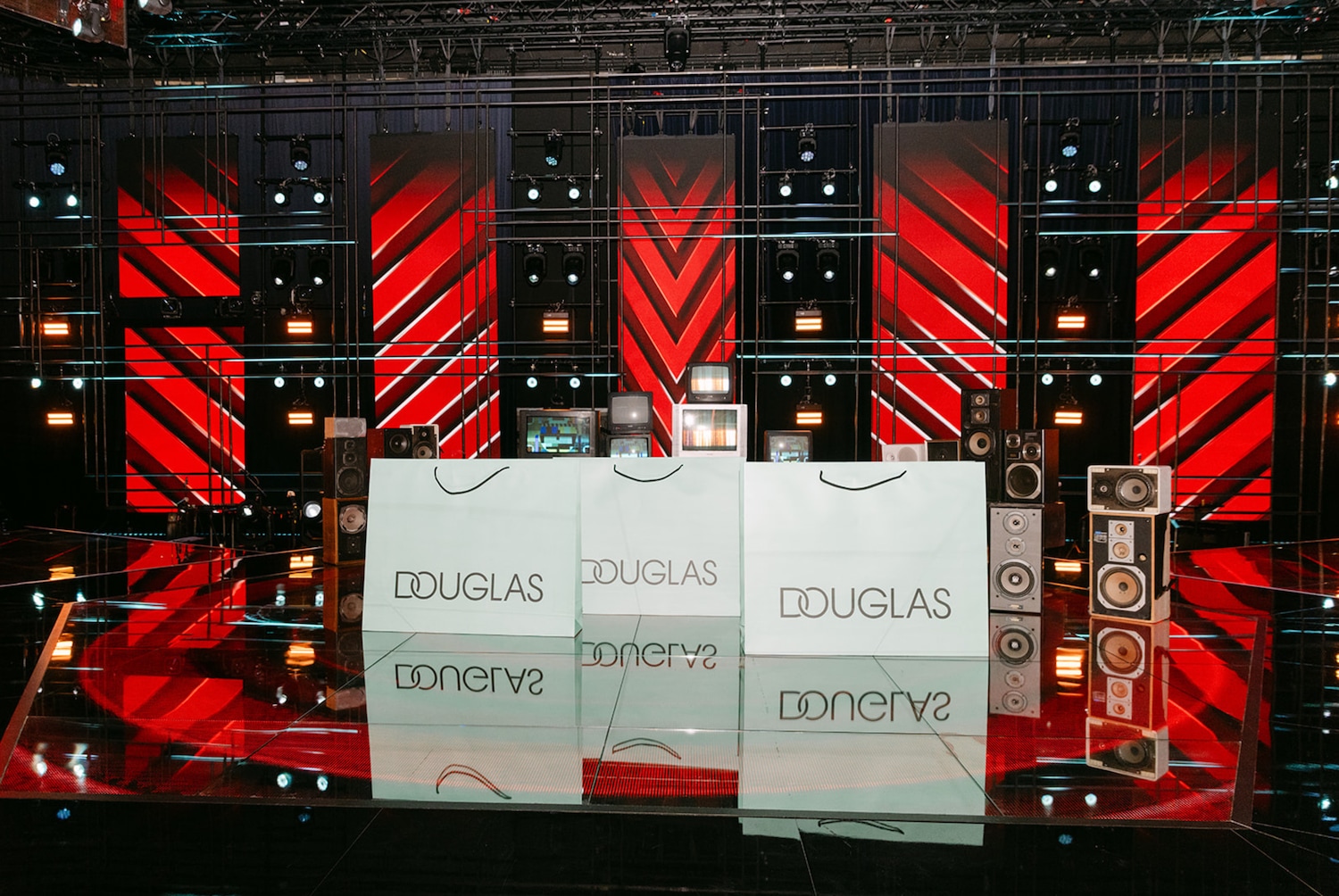 Op een podium staan witte Douglas tassen met daarachter luidsprekers en oude televisies tegen een achtergrond van rode en zwarte strepen.