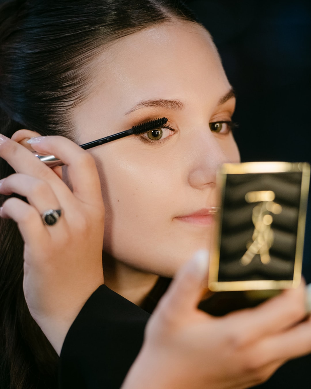 Een jonge vrouw brengt zwarte mascara aan op haar bovenste wimpers, kijkend in een klein, vierkant handspiegeltje met een gouden logo op de zwarte verpakking.
