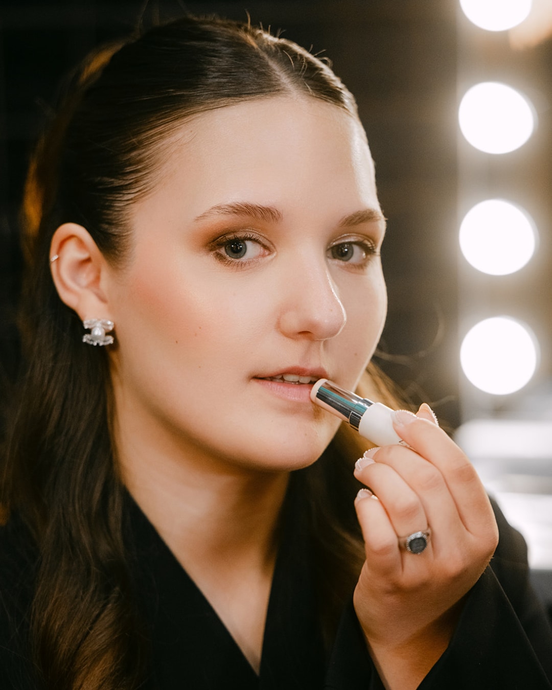 Een jonge vrouw brengt een zilverkleurige lippenbalsem aan, haar haar is opgestoken en ze draagt een zwarte blouse.