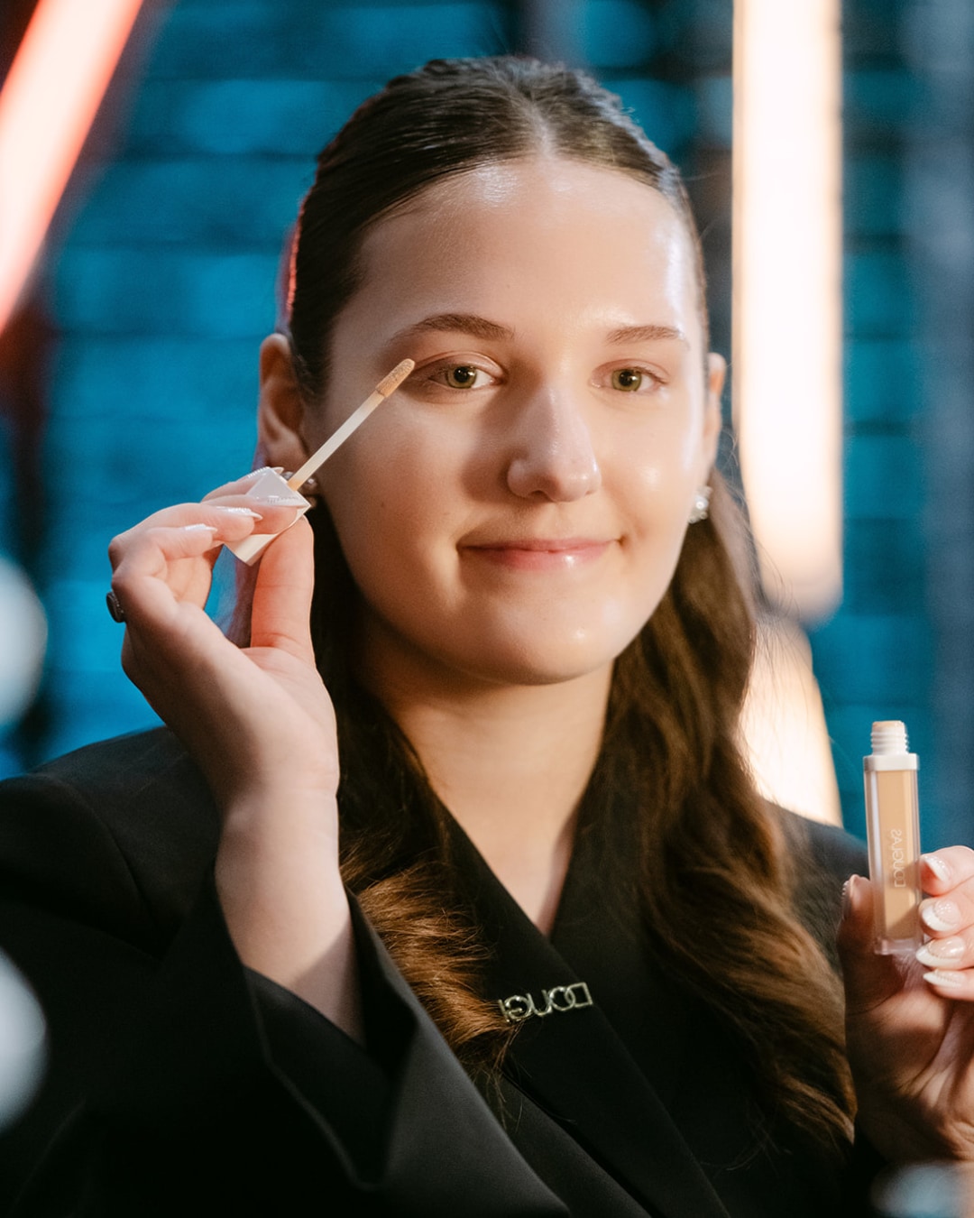 Een jonge vrouw brengt concealer aan op haar ooglid met een applicator, terwijl ze een flesje Douglaskleurige concealer vasthoudt.