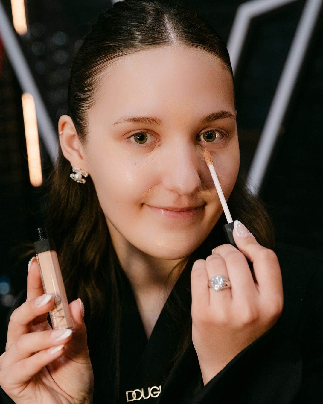 Een vrouw brengt concealer aan onder haar oog met een applicator, terwijl ze een flesje NARS concealer vasthoudt.