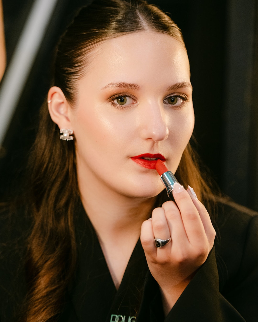 Een vrouw met bruin haar en een zwarte blazer brengt rode lippenstift aan, waardoor haar lippen opvallen.