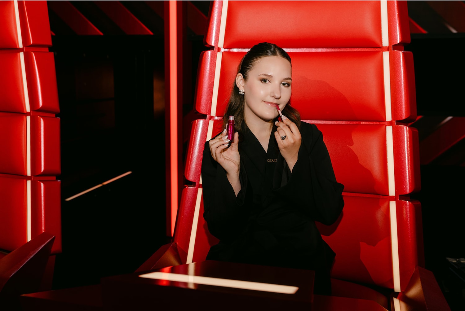 Een jonge vrouw in een zwarte blazer zit in een rode stoel en brengt roze lipgloss aan, waardoor een vleugje glamour aan haar look wordt toegevoegd.