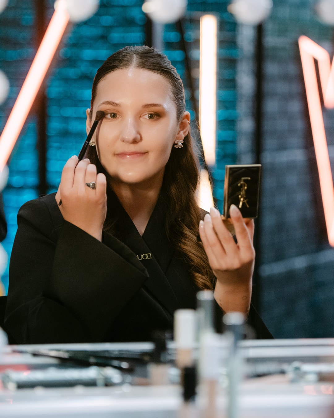 Een vrouw in een zwarte blazer brengt oogschaduw aan met een kwastje, terwijl ze in een klein, goudkleurig YSL-spiegeltje kijkt.