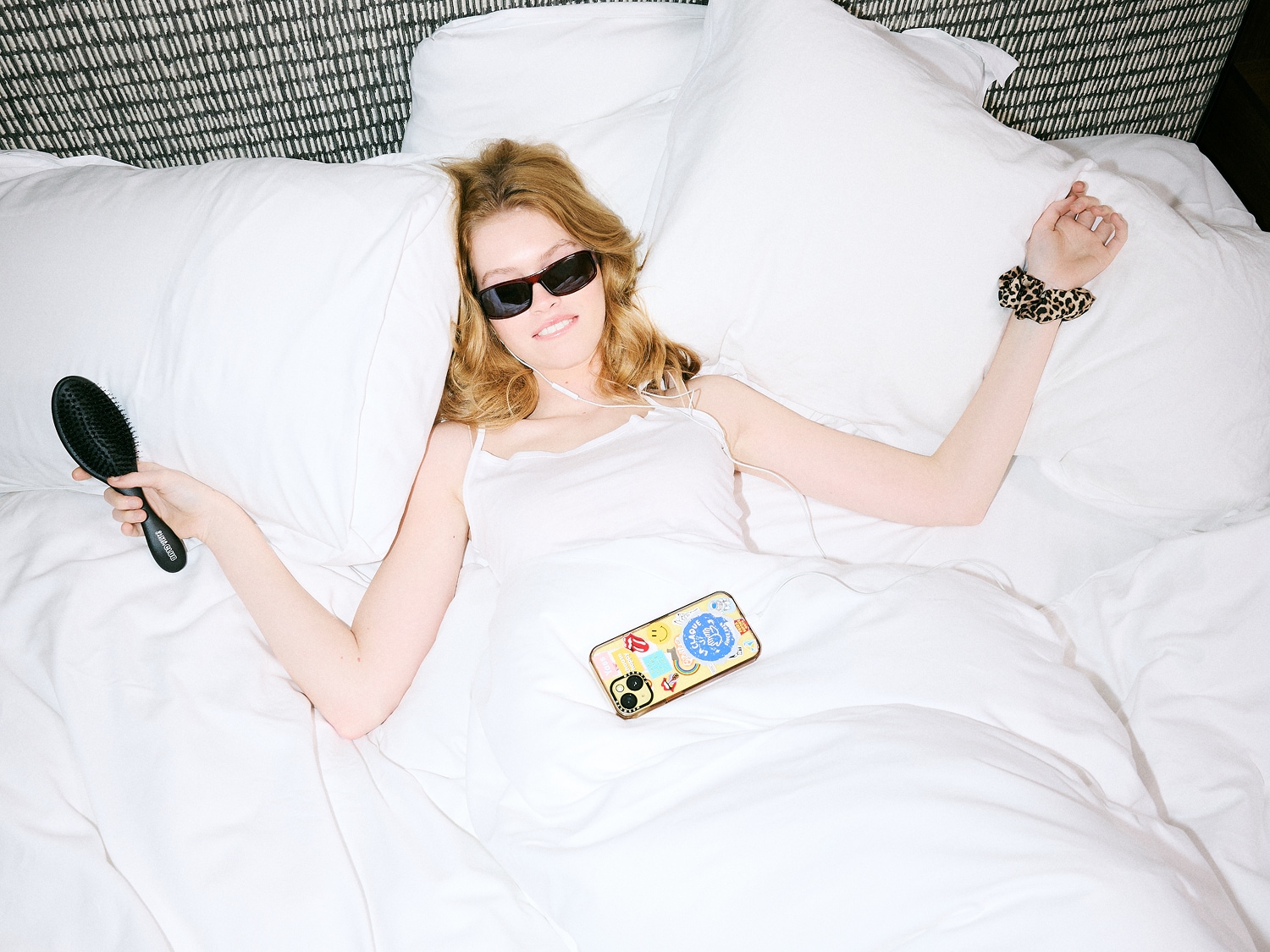 Une femme blonde, lunettes de soleil, allongée sur un lit blanc, tient une brosse à cheveux noire et porte un élastique à cheveux à motif léopard.
