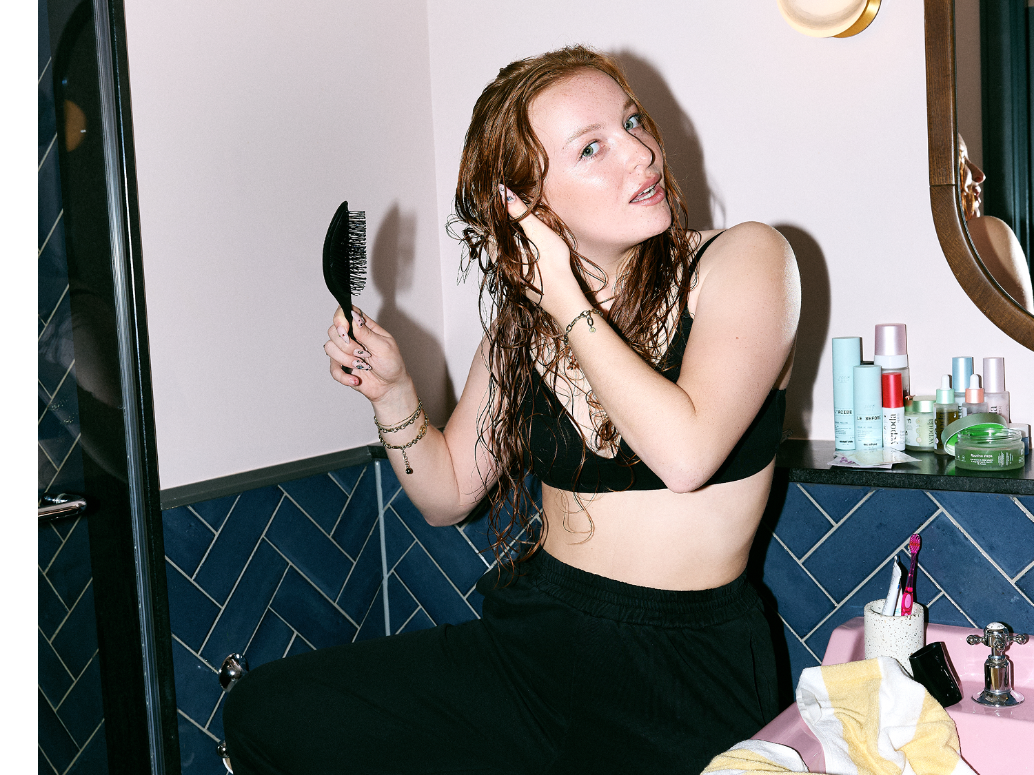Une femme rousse, assise sur le bord d'un lavabo bleu et rose, se brosse les cheveux mouillés avec une brosse noire, portant un soutien-gorge noir et un pantalon de survêtement noir.