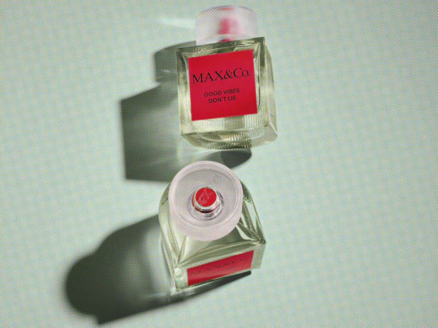 Dwa flakony perfum Max&Co. z przezroczystego szkła z czerwoną etykietą z napisem „GOOD VIBES DON'T LIE” na jasnoniebieskim tle.