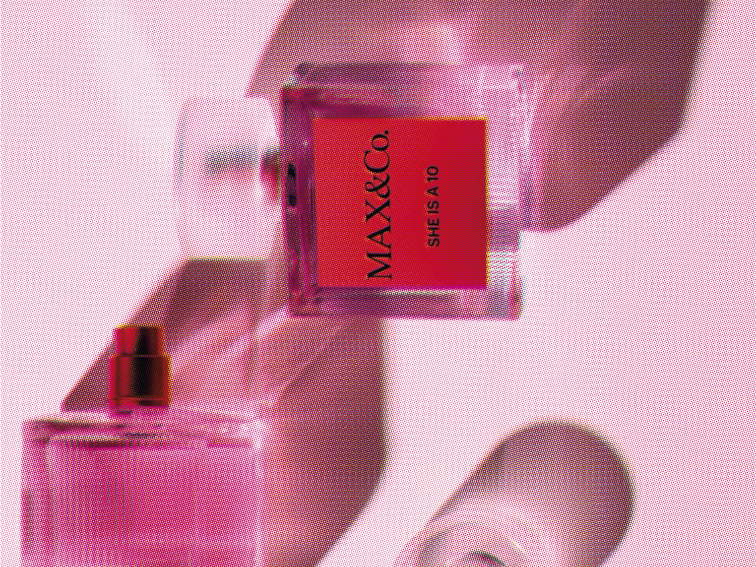 Dwa flakony perfum MAX&Co. SHE IS A 10 na różowym tle, jeden z czerwoną etykietą i otwartym korkiem, drugi z metalowym atomizerem.