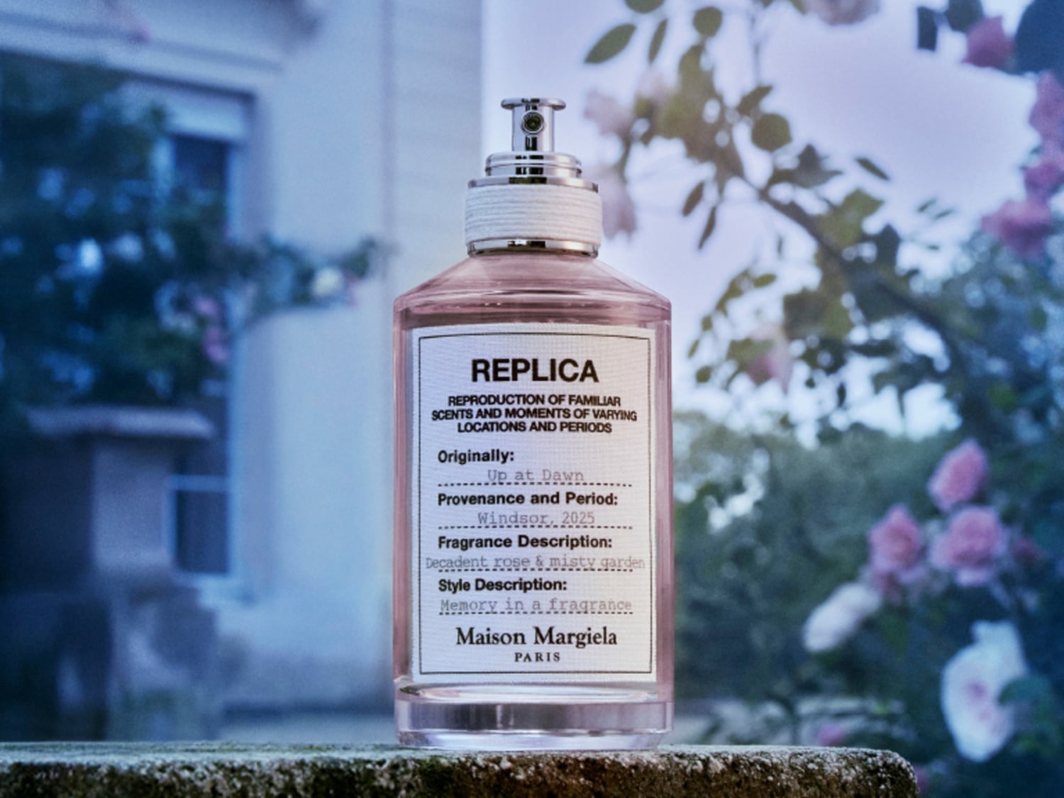 Priehľadný flakón parfému Replica Up at Dawn od Maison Margiela s ružovou tekutinou, etiketou s čiernym písmom a strieborným vrchnákom evokuje vôňu dekadentnej ruže v hmle záhrady.