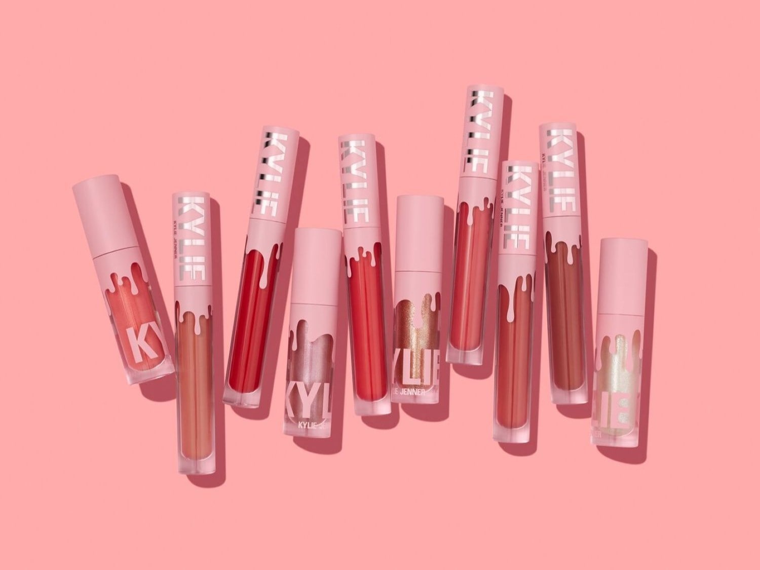 Una gamma di lucidalabbra Kylie Cosmetics in varie tonalità nude e rosate, confezionati in tubi rosa con il logo che cola, su sfondo rosa.