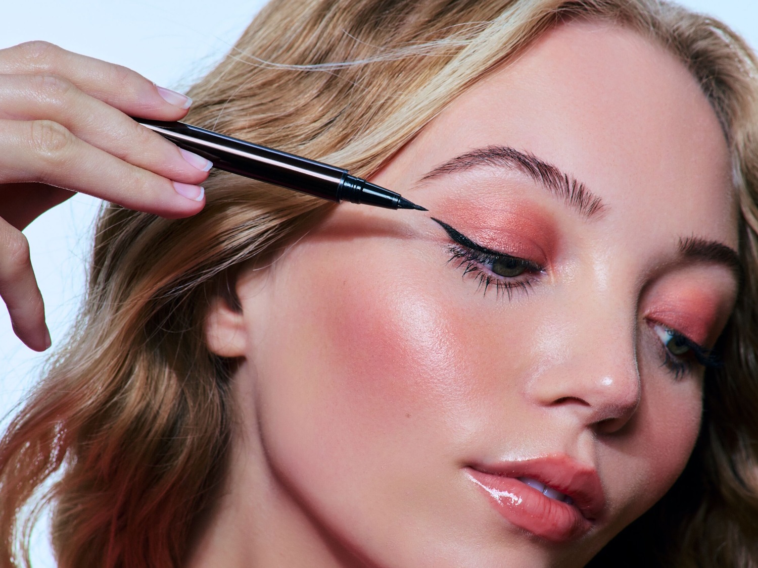 Prim-plan cu o femeie aplicând eyeliner negru cu un aplicator subțire, accentuându-și ochii cu o linie precisă și un machiaj roz delicat.