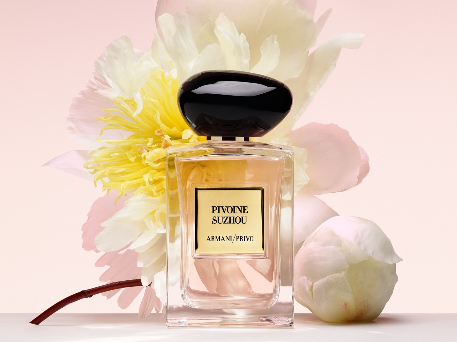 Een heldere glazen fles Armani/Privé Pivoine Suzhou parfum met een zwarte dop staat voor een zachte roze achtergrond met witte en gele bloemen.