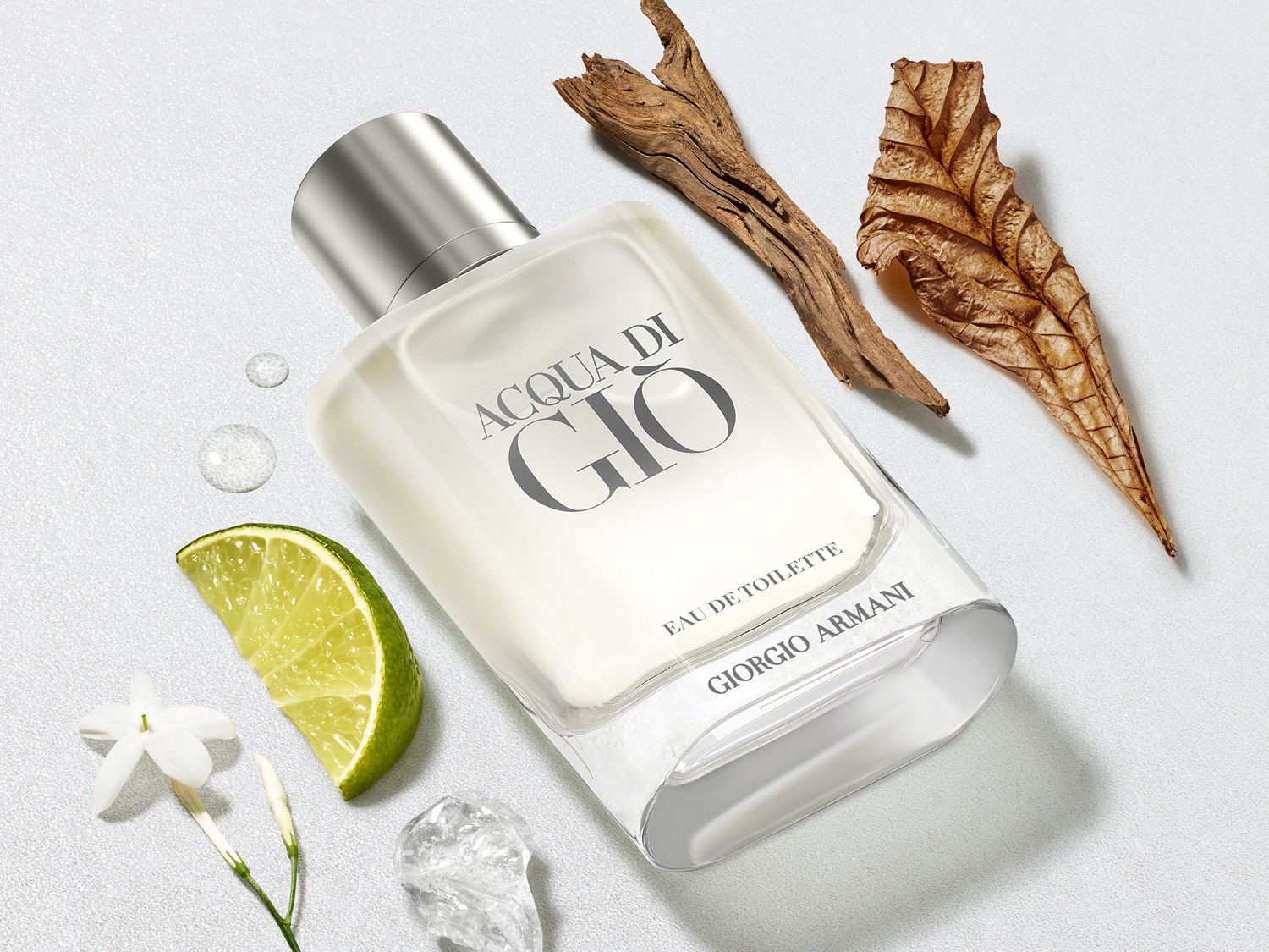 De heldere glazen flacon Acqua di Giò Eau de Toilette van Giorgio Armani, omringd door een schijfje limoen, witte bloemen, stukken drijfhout en een gedroogd blad, straalt een frisse en natuurlijke geur uit.