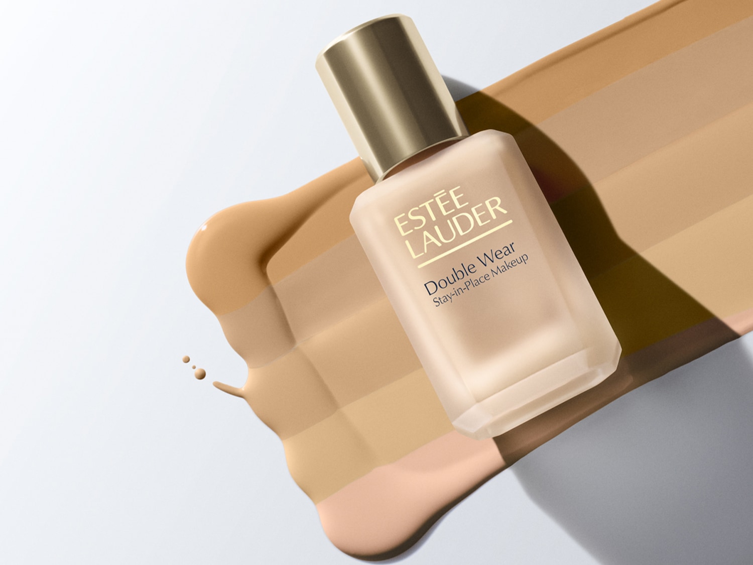 Estée Lauder Double Wear Stay-in-Place Makeup v matné skleněné lahvičce se zlatým víčkem, ležící vedle vzorků různých odstínů make-upu.