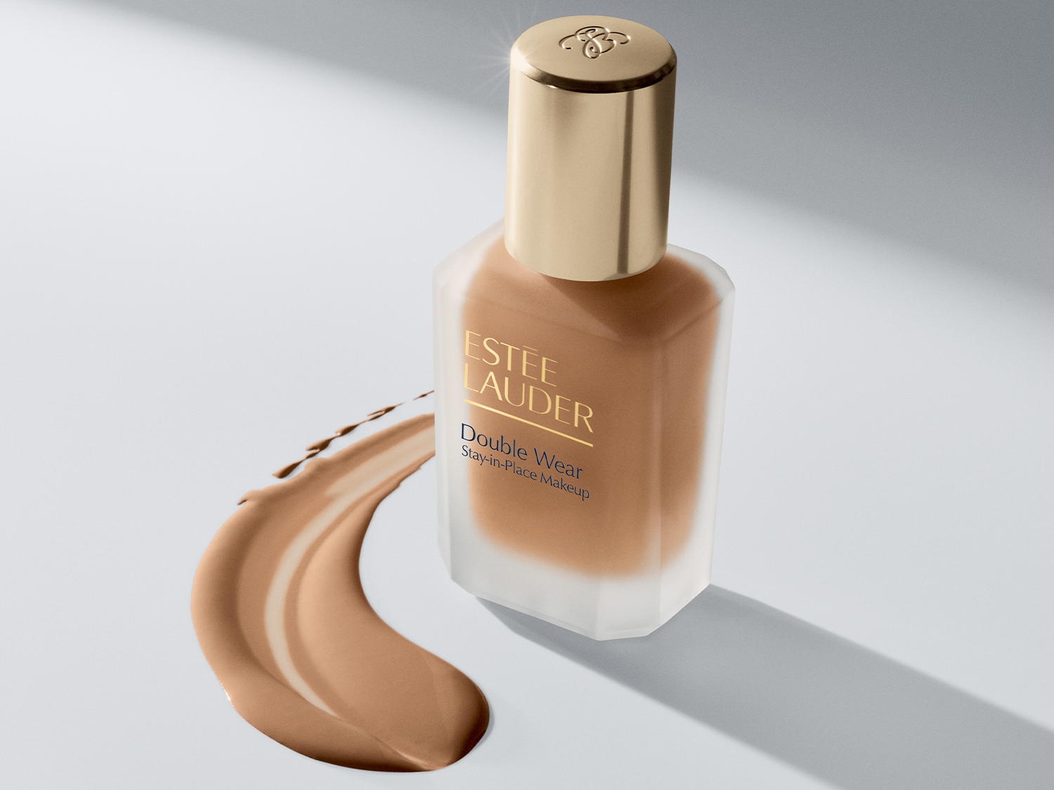 Estée Lauder Double Wear Stay-in-Place Makeup v průhledné skleněné lahvičce s béžovým make-upem a zlatým víčkem, vedle níž je nanesená vrstva make-upu.