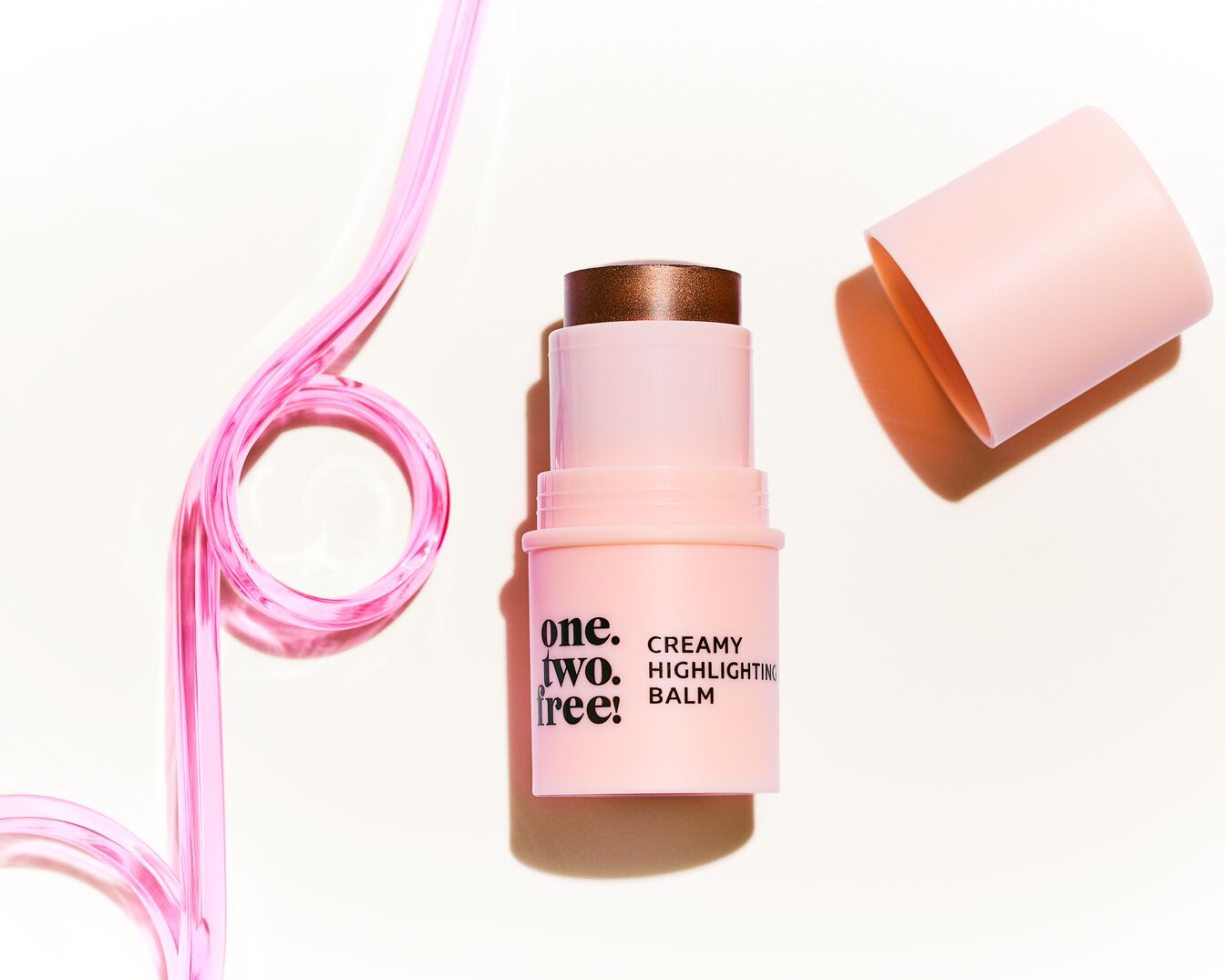 Un baume en stick rose clair "one. two. free!" avec un capuchon ouvert à côté et un tube rose en forme de boucle sur un fond blanc.