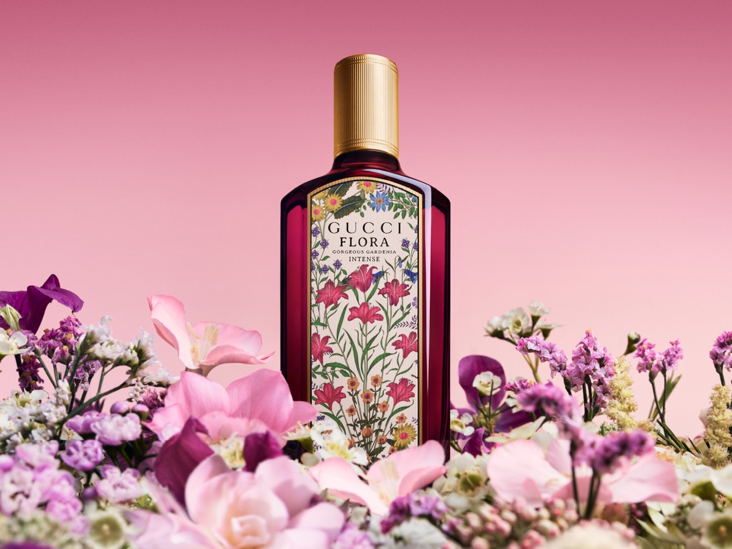 Sticla de parfum Gucci Flora Gorgeous Gardenia Intense, cu capac auriu, este așezată într-un aranjament floral colorat, pe un fundal roz.