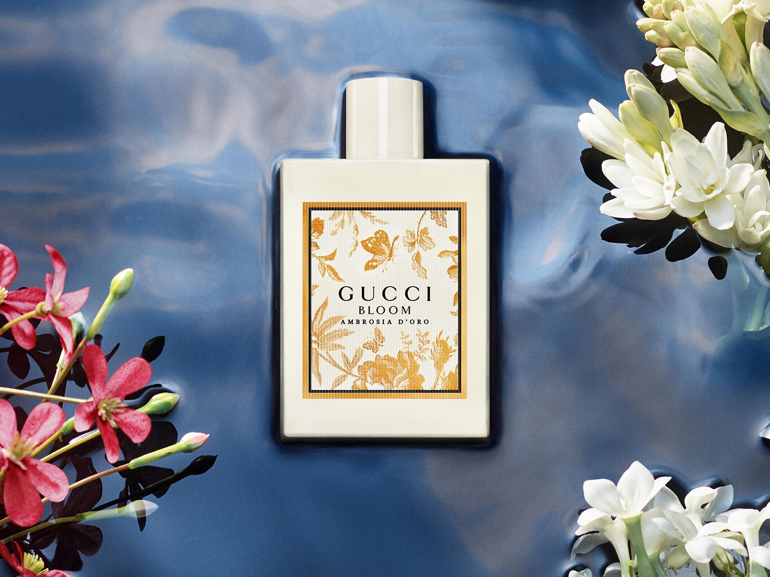 Sticla albă de parfum Gucci Bloom Ambrosia D'oro, cu detalii florale aurii, este așezată într-o apă albastră, înconjurată de flori albe și roșii.