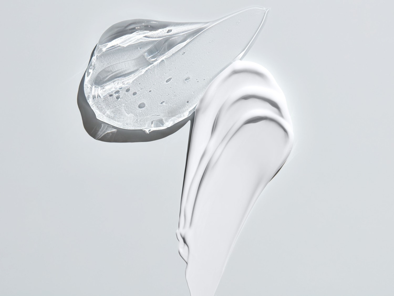 Pe un fundal alb, se află două mostre de produse cosmetice: una este un gel transparent, iar cealaltă este o cremă albă, ambele având o textură netedă și lucioasă.