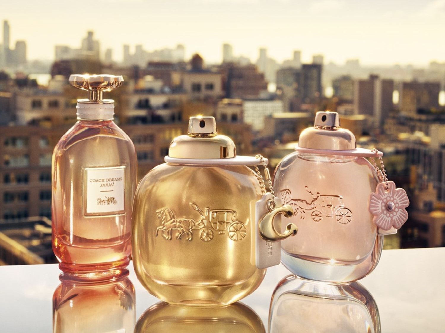 Descoperă Coach Dreams Sunset, un parfum floral-fructat într-o sticlă roz-aurie, alături de Coach Dreams într-o sticlă aurie și Coach Dreams Sunset într-o sticlă transparentă, toate reflectând eleganța brandului.