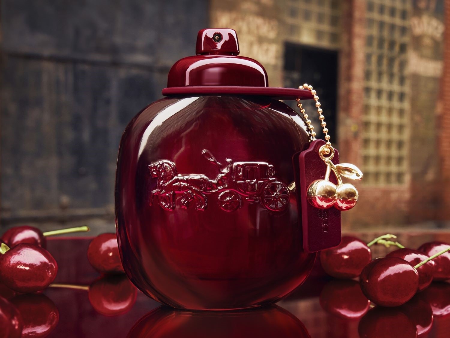 Sticla de parfum Coach Cherry este roșie, lucioasă, cu un logo subtil al trăsurii și un breloc cu cireșe aurii, așezată printre cireșe proaspete.