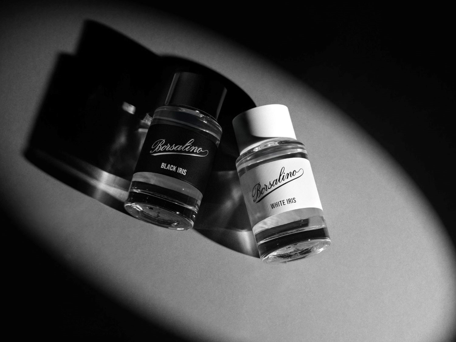 Două sticle de parfum Borsalino, una neagră cu eticheta „Black Iris” și una albă cu eticheta „White Iris”, stau una lângă alta într-o lumină dramatică.