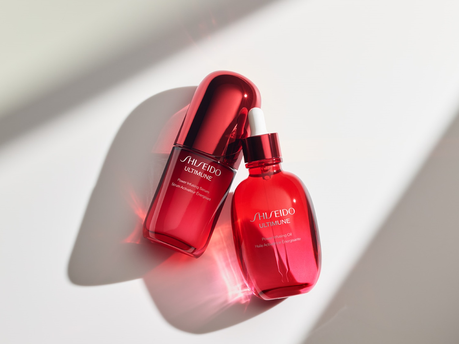 Două flacoane roșii lucioase Shiseido Ultimune, unul serum și celălalt ulei, stau pe o suprafață albă, captând lumina și evidențiind etichetele.