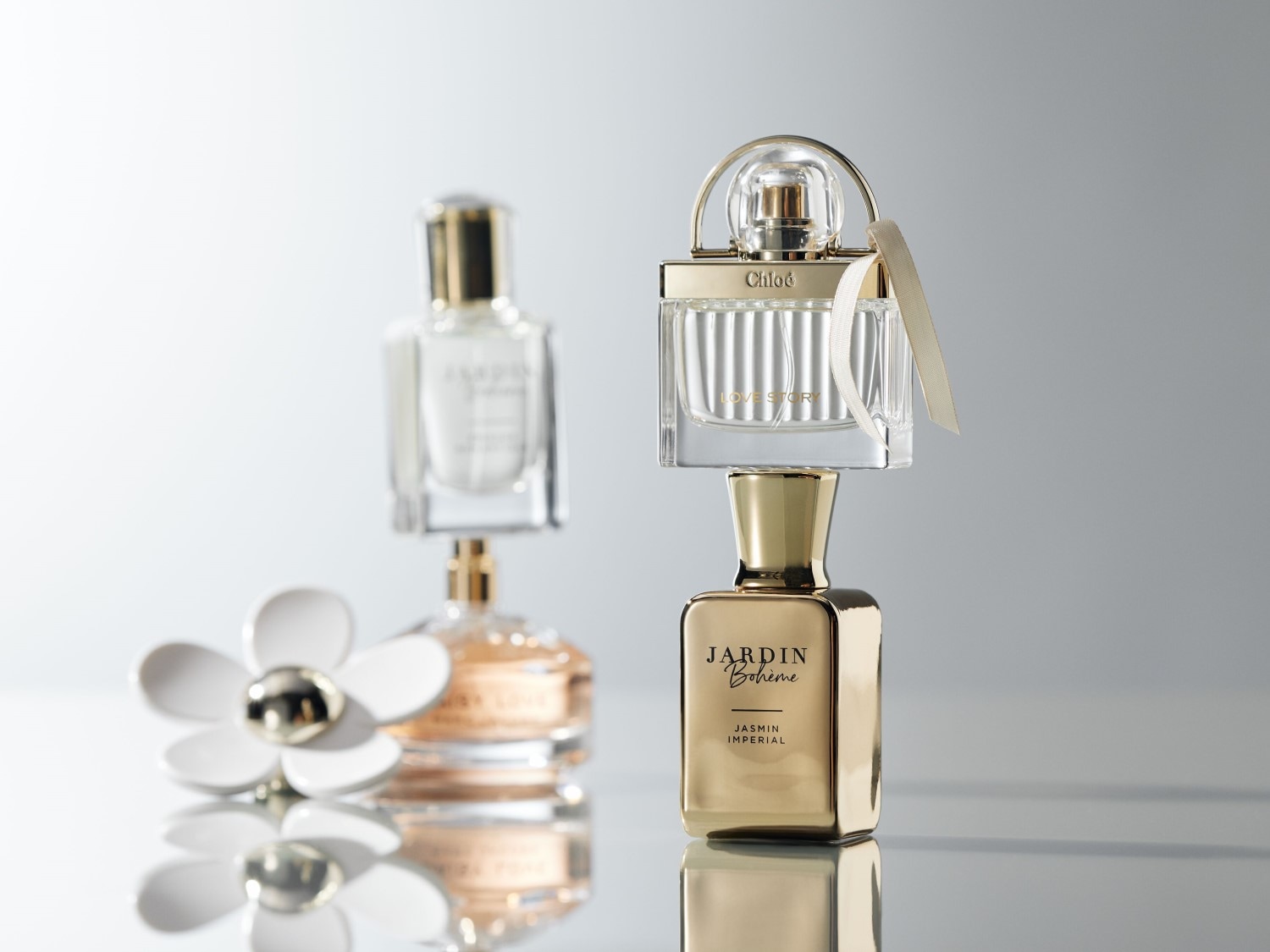 Prezentare cu parfumuri precum Chloé Love Story într-o sticlă transparentă cu fundiță bej, alături de Jardin Bohème Jasmin Imperial într-o sticlă aurie și un parfum Daisy Love într-o sticlă rotundă, toate emanând eleganță.