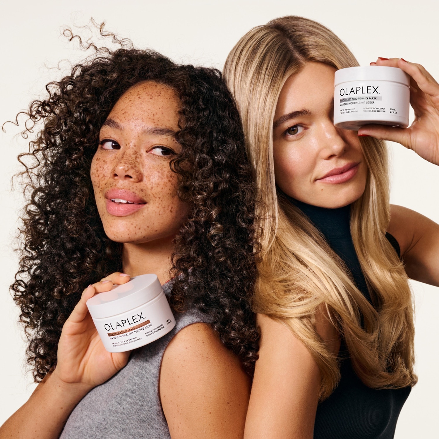 Twee vrouwen, één met krullend haar en de ander met blond haar, houden elk een witte pot Olaplex haarmasker vast, de één voor dik haar en de ander voor fijn haar.