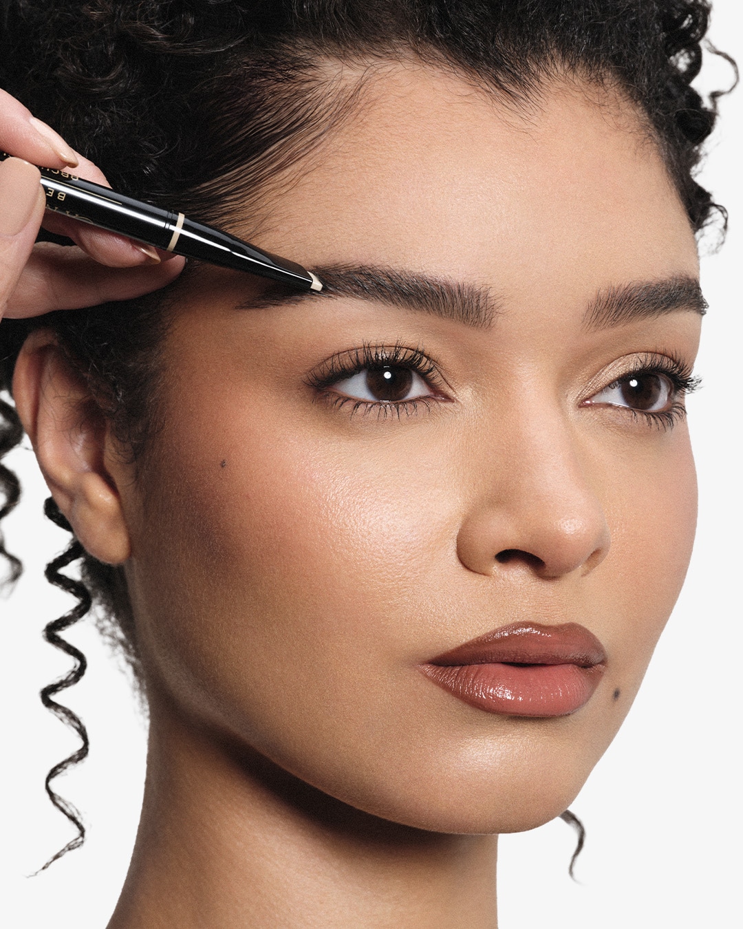 A person uses a black and gold eyebrow pencil to fill in their eyebrow, enhancing their natural beauty.