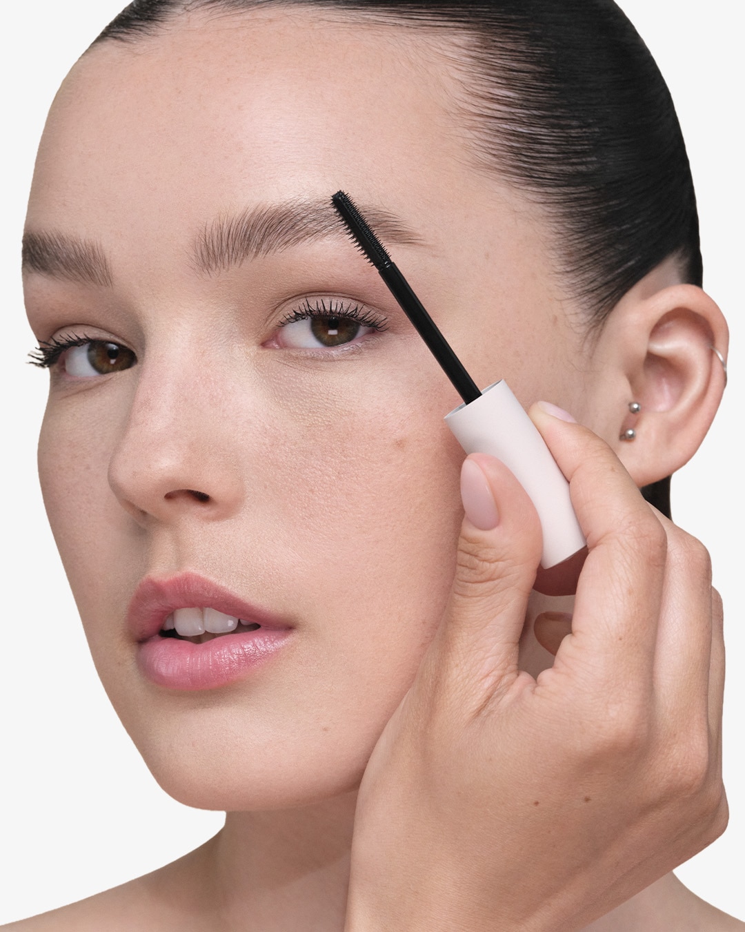 A woman uses a black brush from a white tube to groom her eyebrow, enhancing its shape and definition.