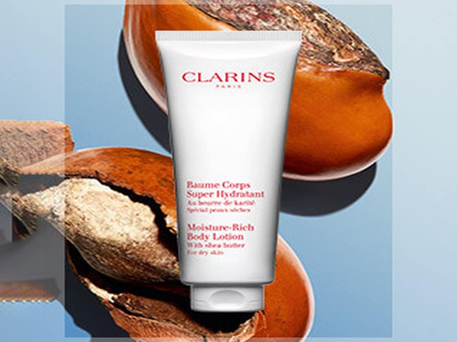 Loțiune de corp Clarins Baume Corps Super Hydratant, într-un tub alb, alături de nuci de shea, oferind hidratare intensă pentru pielea uscată.