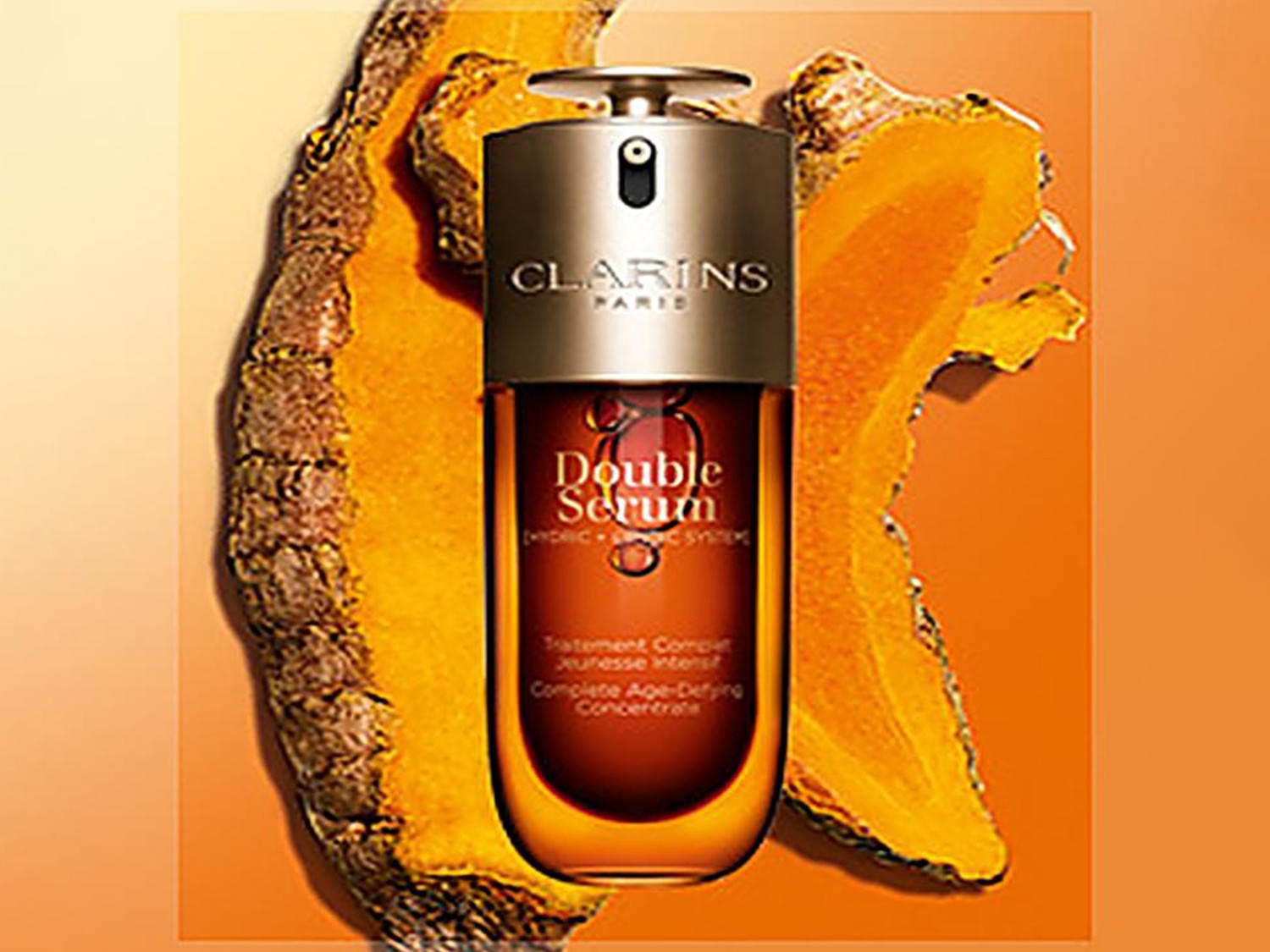 Sticla serului Clarins Double Serum are un capac auriu și o bază maro-portocalie, prezentată alături de o rădăcină de turmeric pe un fundal portocaliu.