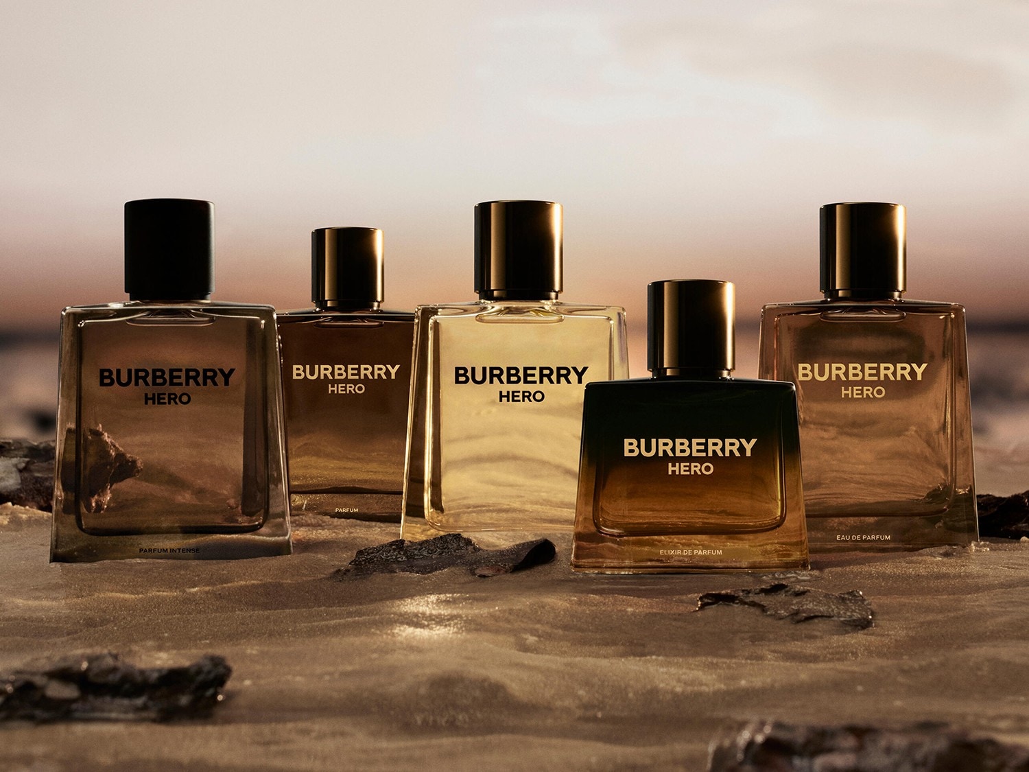 O colecție de parfumuri Burberry Hero în diferite concentrații, inclusiv Parfum Intense, Parfum și Eau de Parfum, prezentate pe un fundal nisipos.