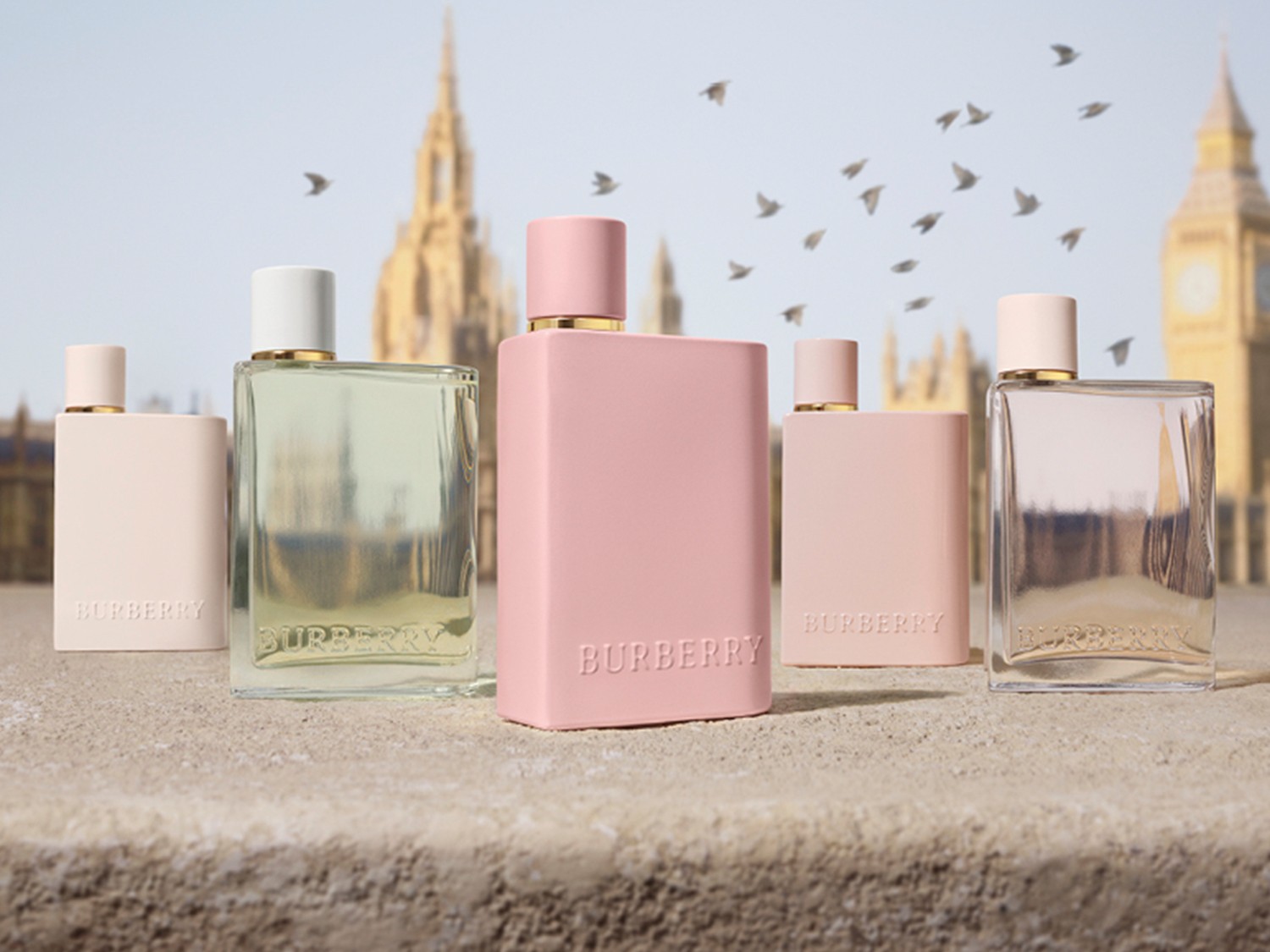O colecție de parfumuri Burberry în nuanțe pastelate, de la roz pudrat la verde pal, prezentate pe un fundal cu repere londoneze și un stol de păsări.